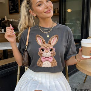 May include: Dark gray t-shirt featuring a crocheted bunny wearing pink glasses and a bow tie. The bunny has large ears and is centered on the shirt. The word "Pepper" is printed on the bottom right of the image.