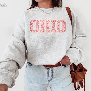 May include: A light grey sweatshirt featuring the word "OHIO" in a coral-coloured, patterned font. The sweatshirt has long sleeves and is worn with light-wash jeans and a brown leather belt. A brown fringed bag is worn over the shoulder.