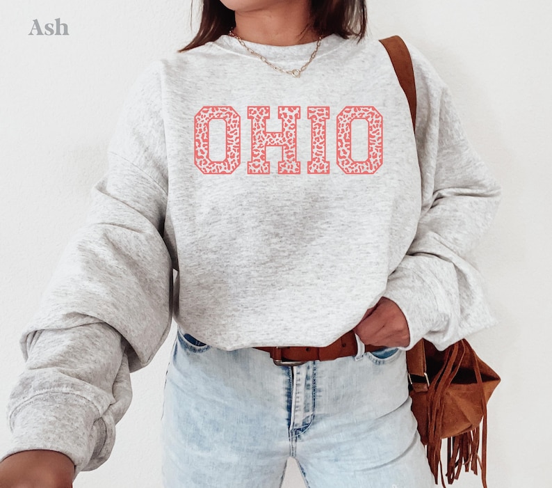 May include: A light grey sweatshirt featuring the word "OHIO" in a coral-coloured, patterned font. The sweatshirt has long sleeves and is worn with light-wash jeans and a brown leather belt. A brown fringed bag is worn over the shoulder.