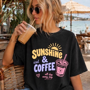Puede incluir: Camiseta negra con un gr&aacute;fico que dice "Sunshine iced & Coffee" en amarillo y morado. El dise&ntilde;o incluye un sol, una taza de caf&eacute; y las palabras "all day every day". La persona de la imagen lleva gafas de sol y bebe de un vaso transparente.