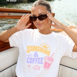 Puede incluir: Camiseta blanca con el texto "SUNSHINE iced & COFFEE all day every day" en amarillo, morado y rosa. El gr&aacute;fico incluye un sol, una taza de caf&eacute; y un coraz&oacute;n. La persona lleva gafas de sol.
