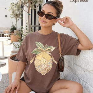 May include: Brown t-shirt with a lemon graphic. The lemon design features a patchwork pattern in yellow, green, and floral prints, with a green gingham leaf and a yellow ribbon. The word "Espresso" is printed in the upper right corner.