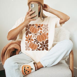 May include: Ivory-colored t-shirt featuring a fall-themed design with pumpkins and flowers in shades of brown and orange. The shirt is paired with gray sweatpants and a matching beanie. The word "Ivory" is printed at the top.