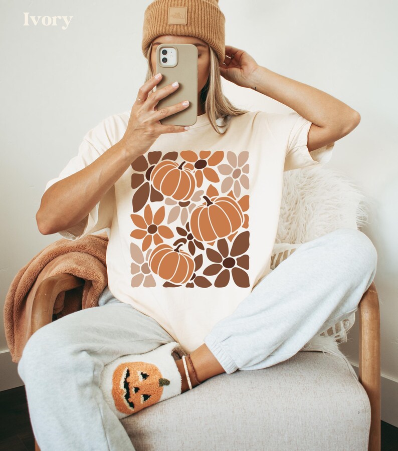 May include: Ivory-colored t-shirt featuring a fall-themed design with pumpkins and flowers in shades of brown and orange. The shirt is paired with gray sweatpants and a matching beanie. The word "Ivory" is printed at the top.