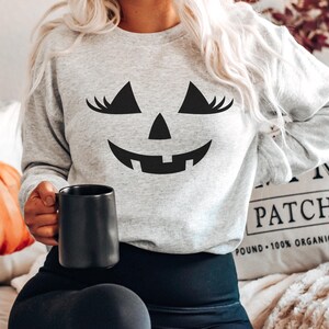 May include: A light grey crewneck sweatshirt featuring a black graphic of a jack-o'-lantern face with long eyelashes. The sweatshirt has long sleeves and a ribbed neckline. The graphic is centred on the chest, depicting triangular eyes with lashes, a triangular nose, and a wide, toothy grin.