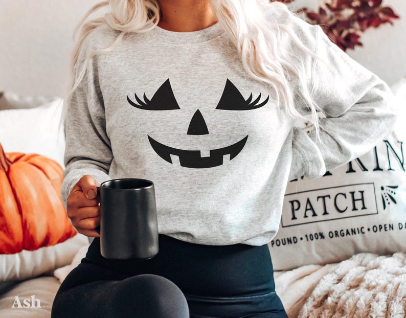 May include: A light grey crewneck sweatshirt featuring a black graphic of a jack-o'-lantern face with long eyelashes. The sweatshirt has long sleeves and a ribbed neckline. The graphic is centred on the chest, depicting triangular eyes with lashes, a triangular nose, and a wide, toothy grin.