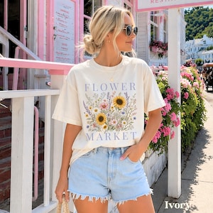 May include: An ivory short-sleeved t-shirt featuring a graphic print of a colorful wildflower bouquet with sunflowers, lavender, and daisies. The text FLOWER MARKET is printed in blue above and below the floral illustration on the front.