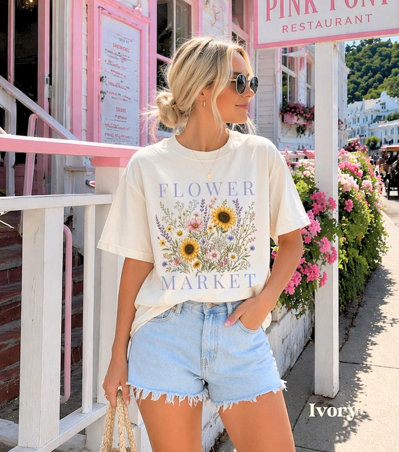 May include: An ivory short-sleeved t-shirt featuring a graphic print of a colorful wildflower bouquet with sunflowers, lavender, and daisies. The text FLOWER MARKET is printed in blue above and below the floral illustration on the front.