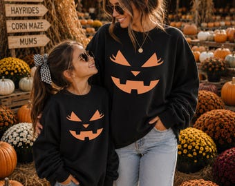 May include: Two black crewneck sweatshirts featuring a large, orange, stylized jack-o'-lantern face graphic on the front. The design includes triangular eyes with eyelashes, a small triangular nose, and a wide, jagged, toothy grin.
