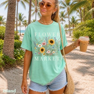 May include: A mint green short-sleeve t-shirt featuring a graphic print of various wildflowers and sunflowers with the text "FLOWER MARKET" in white lettering. The shirt has a crew neckline and a relaxed fit, paired with light blue denim shorts.