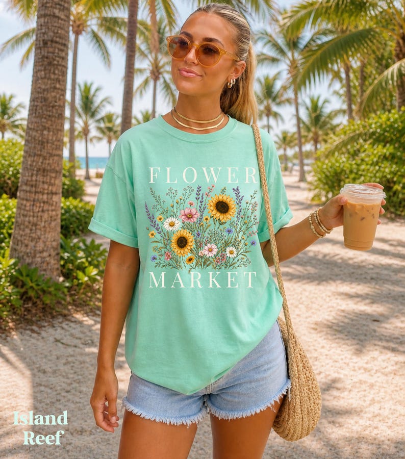 May include: A mint green short-sleeve t-shirt featuring a graphic print of various wildflowers and sunflowers with the text "FLOWER MARKET" in white lettering. The shirt has a crew neckline and a relaxed fit, paired with light blue denim shorts.