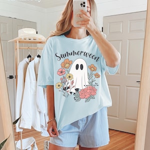 May include: Light blue t-shirt with the word "Summerween" and a ghost graphic surrounded by flowers. The shirt is part of a summer collection and is made of a soft material. The model is wearing blue and white striped shorts.