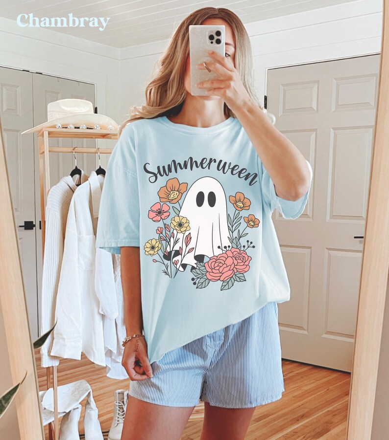 May include: Light blue t-shirt with the word "Summerween" and a ghost graphic surrounded by flowers. The shirt is part of a summer collection and is made of a soft material. The model is wearing blue and white striped shorts.