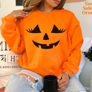 May include: A neon safety orange long-sleeve sweatshirt featuring a black graphic of a jack-o'-lantern face with eyelashes. The text Neon Safety Orange is printed in the upper right corner. The garment has a crew neckline and long sleeves.