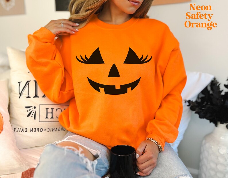 May include: A neon safety orange long-sleeve sweatshirt featuring a black graphic of a jack-o'-lantern face with eyelashes. The text Neon Safety Orange is printed in the upper right corner. The garment has a crew neckline and long sleeves.