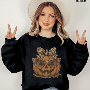 Op de afbeelding: Een zwart sweatshirt met ronde hals en een afbeelding van een oranje pompoen met een zonnebril met luipaardprint en een bijpassende strik. De pompoen rust op een klein hoopje bruine herfstbladeren.