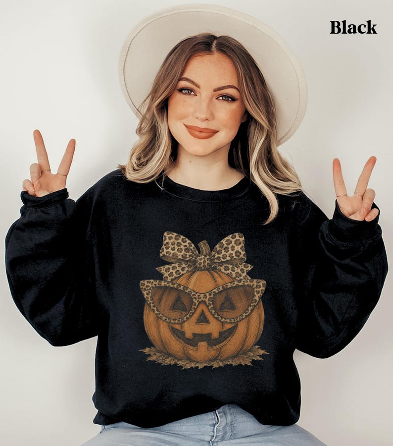 Op de afbeelding: Een zwart sweatshirt met ronde hals en een afbeelding van een oranje pompoen met een zonnebril met luipaardprint en een bijpassende strik. De pompoen rust op een klein hoopje bruine herfstbladeren.