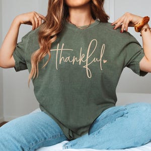 May include: A moss green t-shirt with the word "thankful" in a cursive font. The shirt is paired with light blue jeans. The word "Moss" is printed in the bottom right corner.