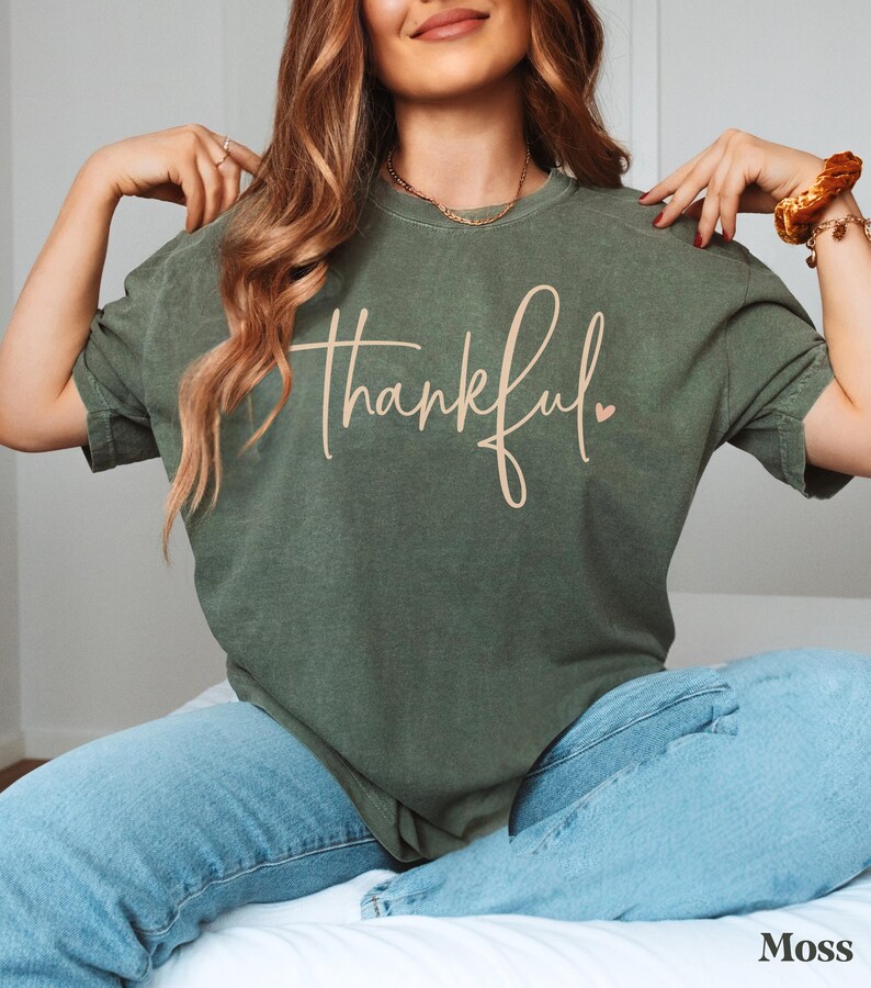 May include: A moss green t-shirt with the word "thankful" in a cursive font. The shirt is paired with light blue jeans. The word "Moss" is printed in the bottom right corner.