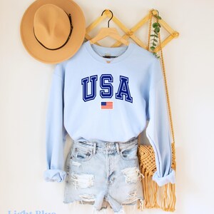 Pu&ograve; includere: Felpa celeste con la scritta "USA" in lettere blu scuro e un piccolo grafico della bandiera americana.