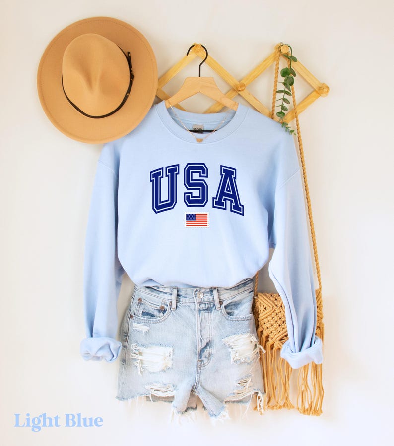 Pu&ograve; includere: Felpa celeste con la scritta "USA" in lettere blu scuro e un piccolo grafico della bandiera americana.