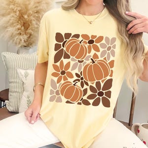 May include: A light yellow t-shirt featuring a fall-themed design. The graphic includes three pumpkins and floral elements in shades of brown and tan. The word "Butter" is printed at the bottom right.