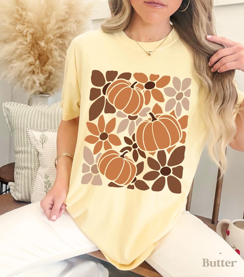 May include: A light yellow t-shirt featuring a fall-themed design. The graphic includes three pumpkins and floral elements in shades of brown and tan. The word "Butter" is printed at the bottom right.
