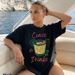 May include: Black t-shirt with a colorful graphic of a cocktail, a lime slice, and a chili pepper. The text "Cinco de Drinko" is printed on the front. The word "Black" is printed at the bottom of the shirt.