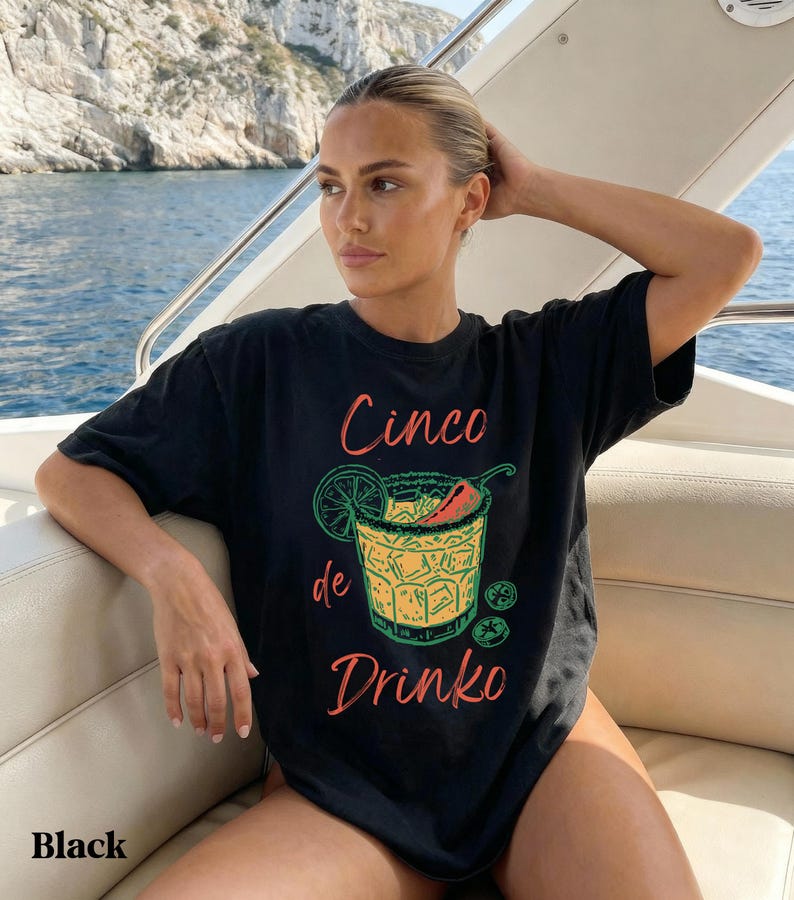 May include: Black t-shirt with a colorful graphic of a cocktail, a lime slice, and a chili pepper. The text "Cinco de Drinko" is printed on the front. The word "Black" is printed at the bottom of the shirt.
