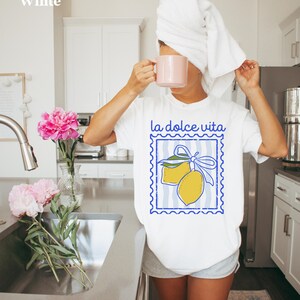 Pu&ograve; includere: T-shirt bianco con grafica di limone blu e giallo e la scritta "la dolce vita". La persona tiene in mano una tazza rosa e indossa un asciugamano bianco sulla testa. Peonie rosa in un vaso.
