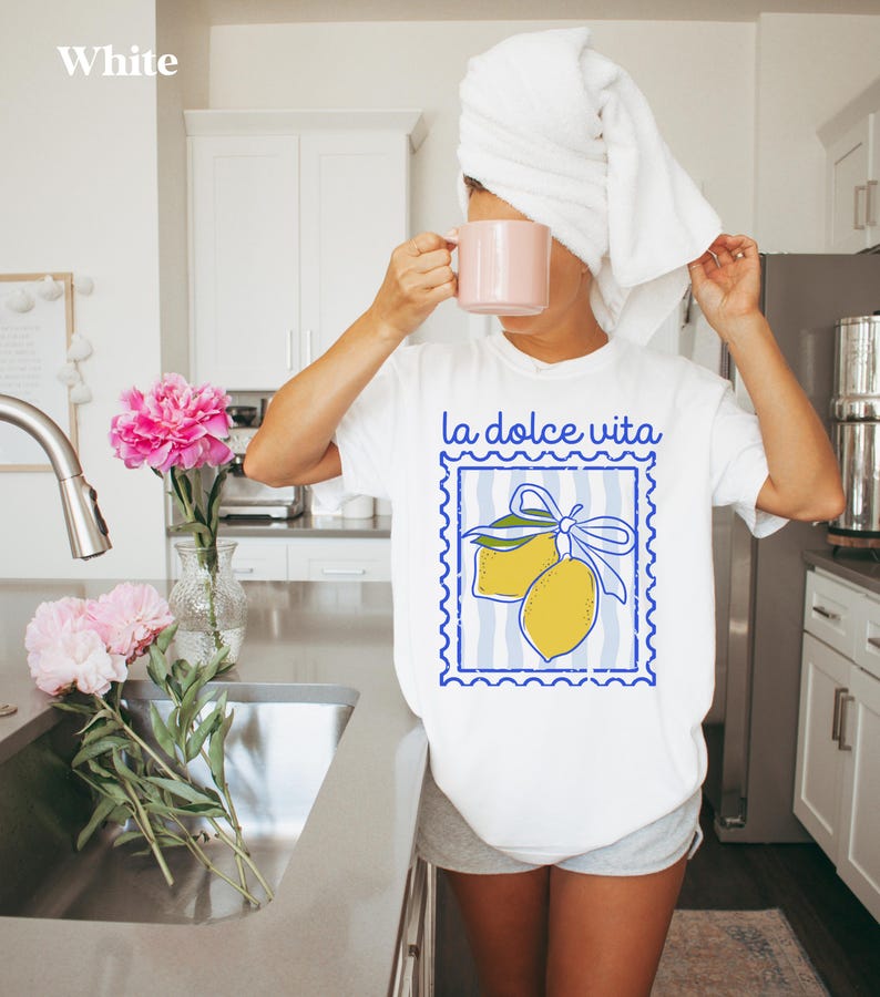 Pu&ograve; includere: T-shirt bianco con grafica di limone blu e giallo e la scritta "la dolce vita". La persona tiene in mano una tazza rosa e indossa un asciugamano bianco sulla testa. Peonie rosa in un vaso.