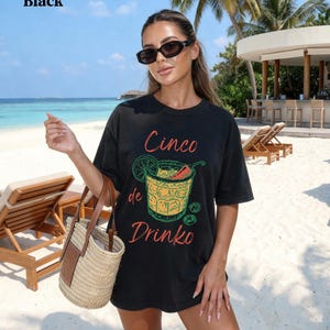 May include: Black t-shirt with the text "Cinco de Drinko" and a graphic of a cocktail. The shirt is worn on a beach with a straw tote bag. The background includes the ocean, beach chairs, and a bar.