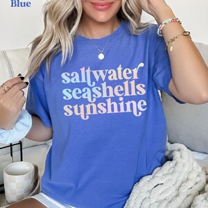 sunshine beach vacation shirt that says saltwater seashells sunshine in a fun summer print on a Comfort Colors shirt in the color flo blue