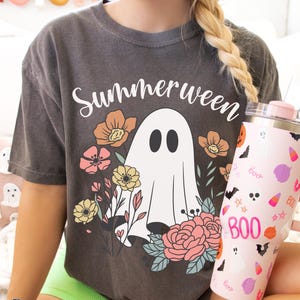 May include: A dark gray t-shirt with the word "Summerween" above a white ghost illustration surrounded by flowers. A pink tumbler with Halloween-themed graphics is held in the hand.