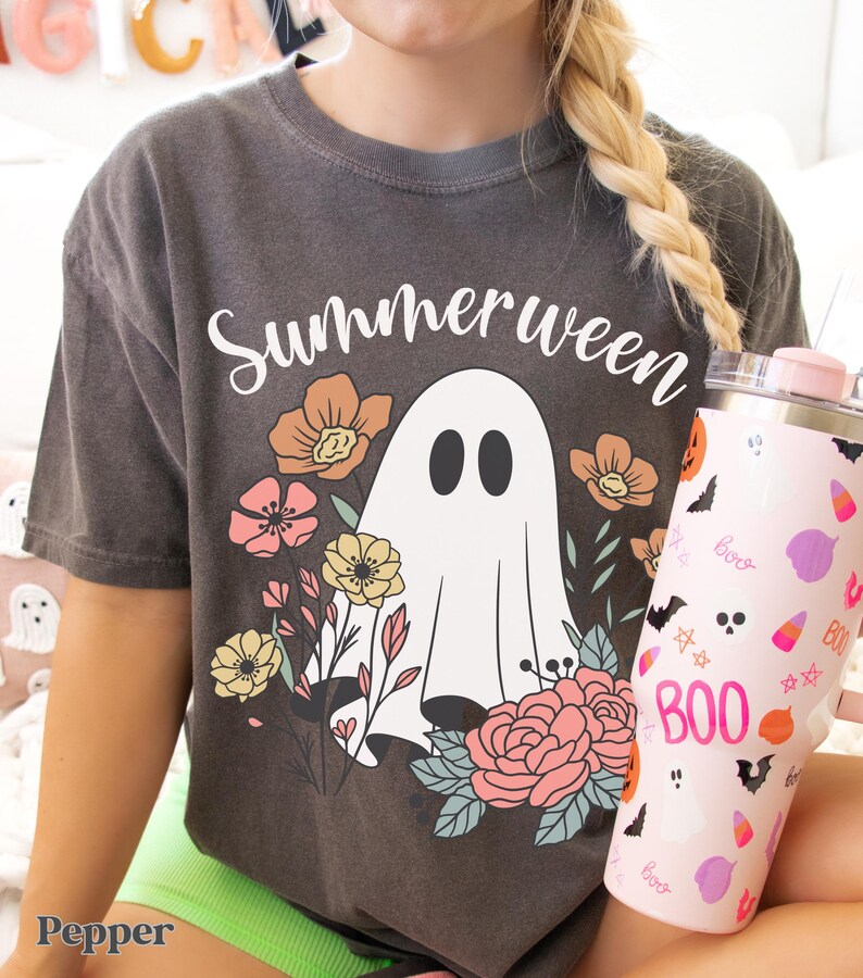 May include: A dark gray t-shirt with the word "Summerween" above a white ghost illustration surrounded by flowers. A pink tumbler with Halloween-themed graphics is held in the hand.
