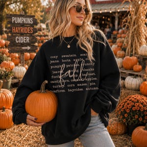 May include: A black long-sleeve sweatshirt featuring white text that lists fall-themed words including cozy sweaters, jeans, boots, apple cider, pumpkin patches, crunchy leaves, giving thanks, football, soup, harvest, candy corn, caramel apples, turkey, corn mazes, pie, bonfires, hayrides, and autumn.