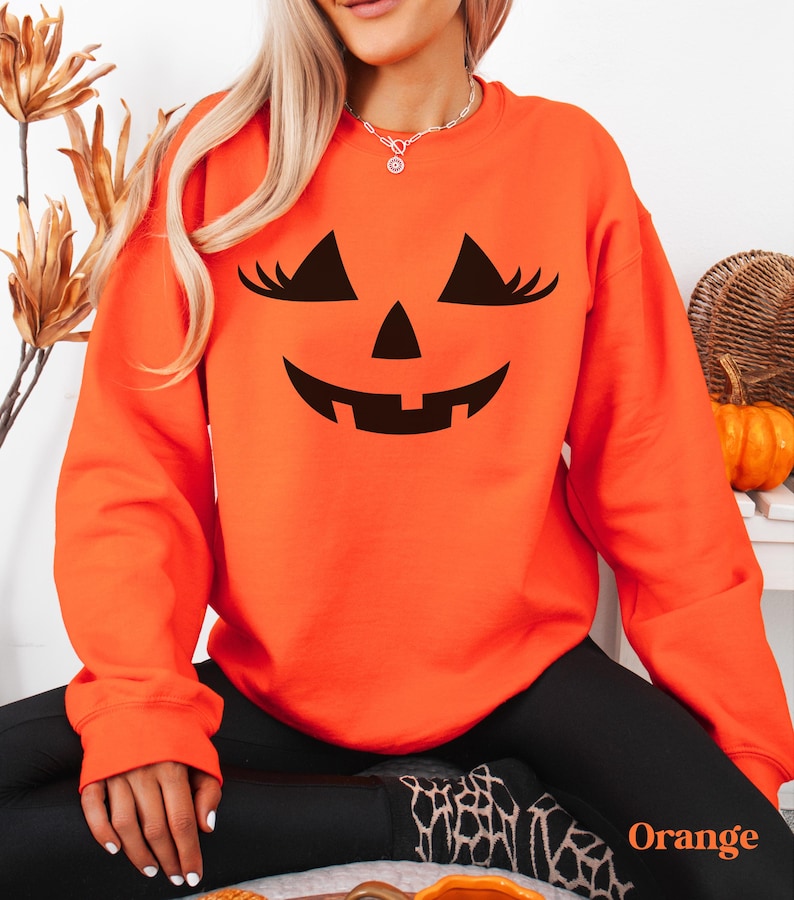 May include: A bright orange long-sleeve sweatshirt featuring a black graphic print of a jack-o'-lantern face with eyelashes on the front. The design includes triangular eyes with long lashes, a triangular nose, and a wide, toothy grin.
