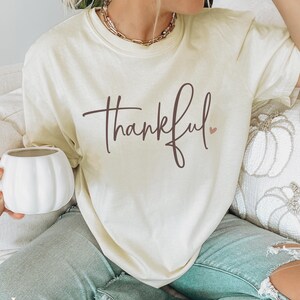 May include: Ivory-colored t-shirt with the word "thankful" in a brown script font and a small pink heart. The shirt is worn with light blue jeans. A white pumpkin-shaped mug is held in the hand.