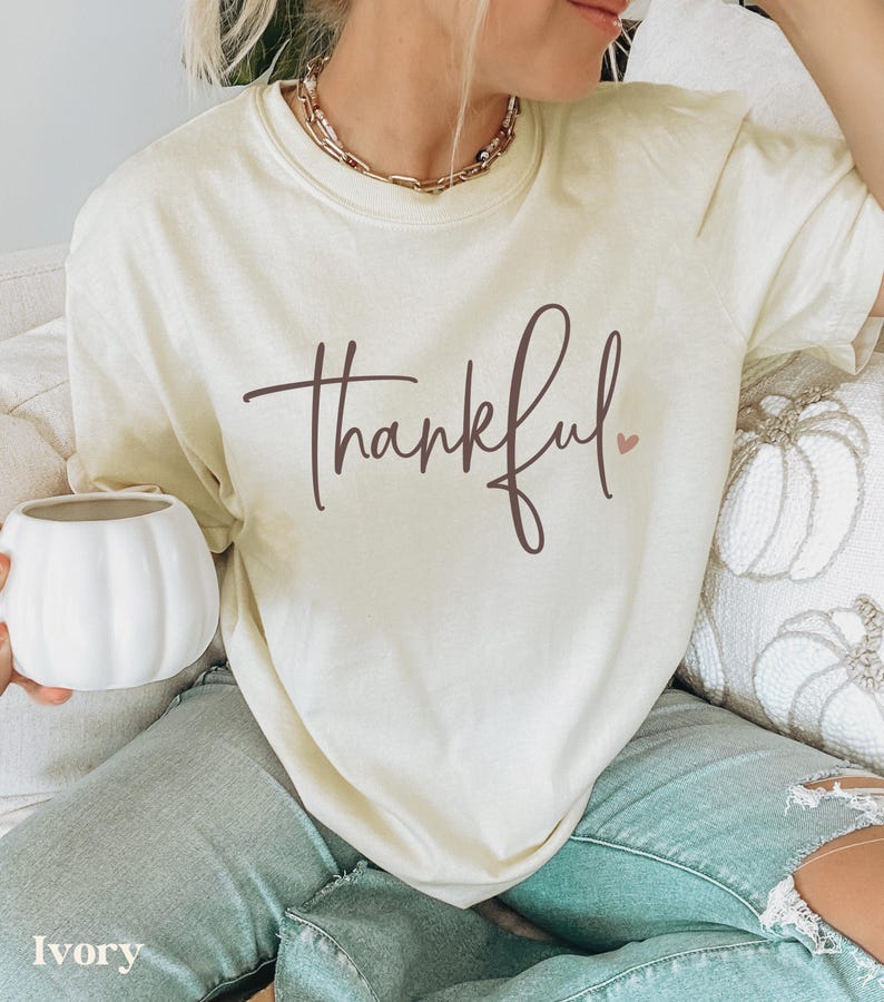 May include: Ivory-colored t-shirt with the word "thankful" in a brown script font and a small pink heart. The shirt is worn with light blue jeans. A white pumpkin-shaped mug is held in the hand.