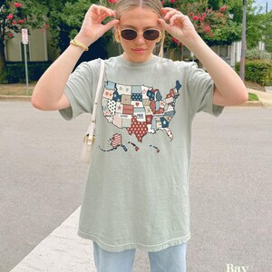 May include: A light green t-shirt with a patchwork design of the United States of America. The patches are red, white, and blue and feature words like "Brave", "Loved", and "Free".
