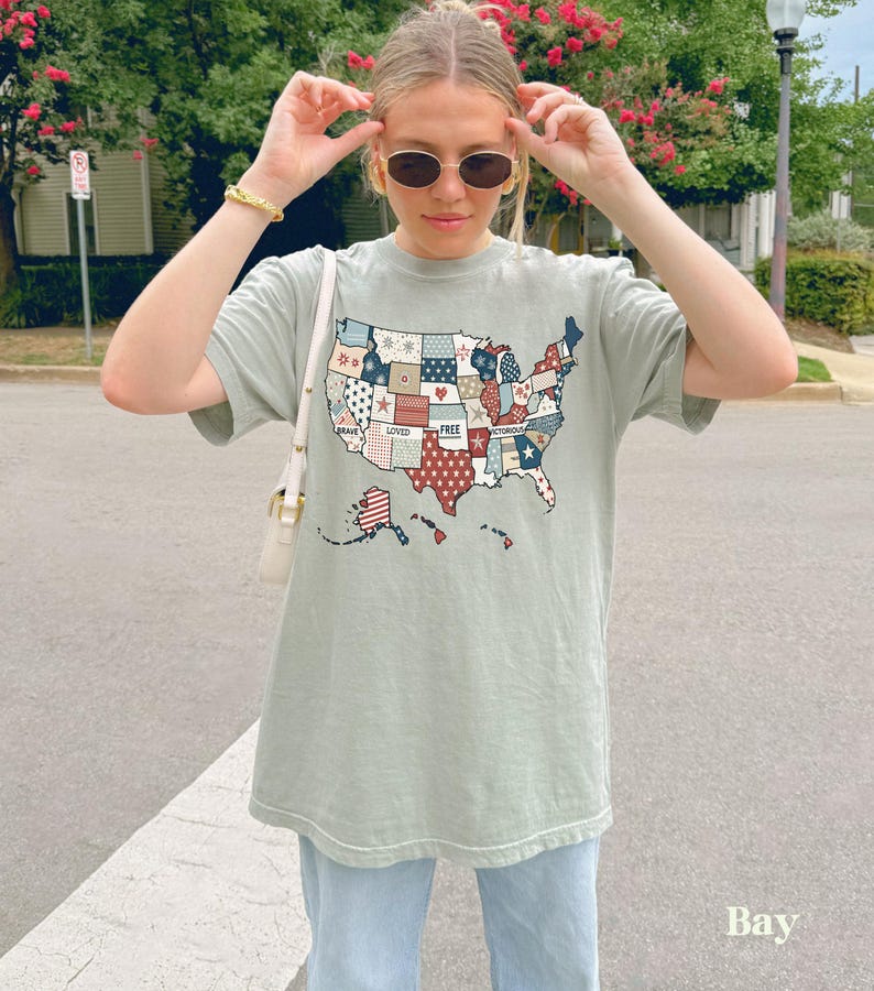 May include: A light green t-shirt with a patchwork design of the United States of America. The patches are red, white, and blue and feature words like "Brave", "Loved", and "Free".