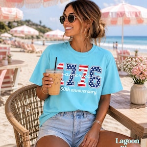 May include: A turquoise t-shirt featuring the text "1776 America" in a patriotic design, celebrating a 50th anniversary. The shirt is paired with denim shorts. The image is taken outdoors on a sunny day.