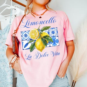 Pu&ograve; includere: Maglietta rosa con una grafica di limoni e piastrelle blu e bianche. Il testo "Limoncello La Dolce Vita" &egrave; stampato sulla maglietta.