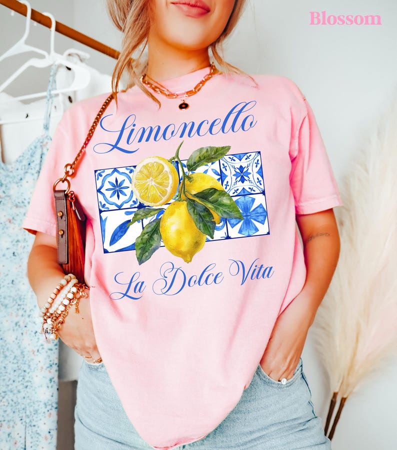 Pu&ograve; includere: Maglietta rosa con una grafica di limoni e piastrelle blu e bianche. Il testo "Limoncello La Dolce Vita" &egrave; stampato sulla maglietta.