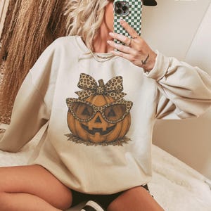 Op de afbeelding: Een zandkleurige sweater met ronde hals en een centrale afbeelding van een oranje pompoen met een zonnebril en een strik met luipaardprint. De pompoen heeft een uitgesneden gezicht en rust op een kleine hoop bruine bladeren.