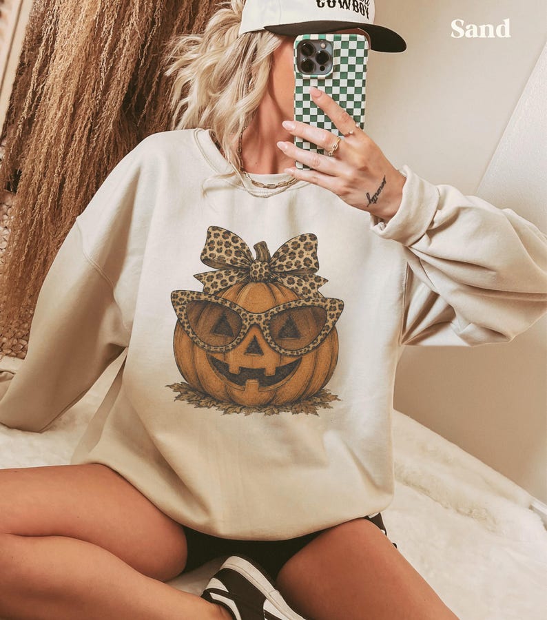 Op de afbeelding: Een zandkleurige sweater met ronde hals en een centrale afbeelding van een oranje pompoen met een zonnebril en een strik met luipaardprint. De pompoen heeft een uitgesneden gezicht en rust op een kleine hoop bruine bladeren.