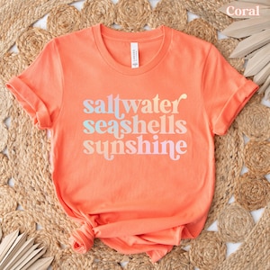 May include: Coral-colored t-shirt with rolled sleeves. The shirt features the words "saltwater seashells sunshine" in a gradient font. The shirt is made of a soft material and is displayed on a woven surface.