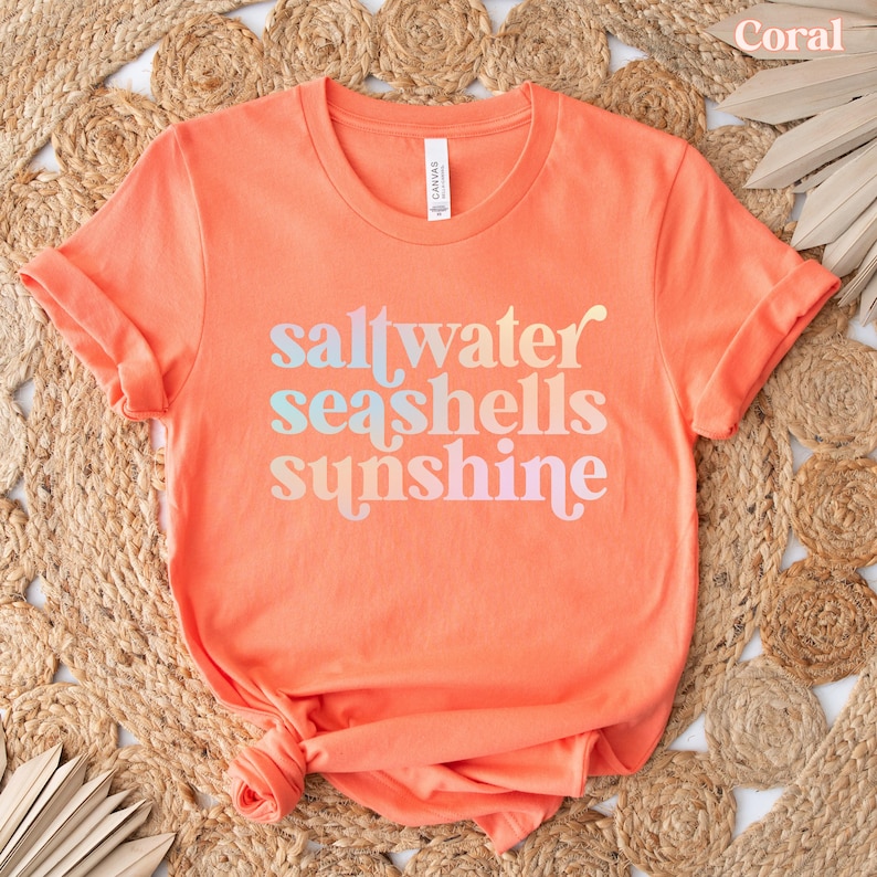 May include: Coral-colored t-shirt with rolled sleeves. The shirt features the words "saltwater seashells sunshine" in a gradient font. The shirt is made of a soft material and is displayed on a woven surface.
