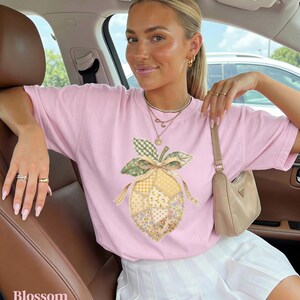 May include: A pale pink t-shirt with a patchwork lemon design, green leaves, and a bow. The word "Blossom" is printed in white at the bottom left. The shirt is paired with a white skirt.