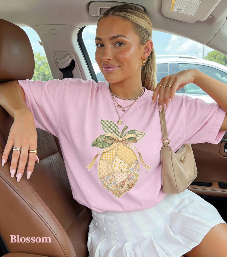 May include: A pale pink t-shirt with a patchwork lemon design, green leaves, and a bow. The word "Blossom" is printed in white at the bottom left. The shirt is paired with a white skirt.